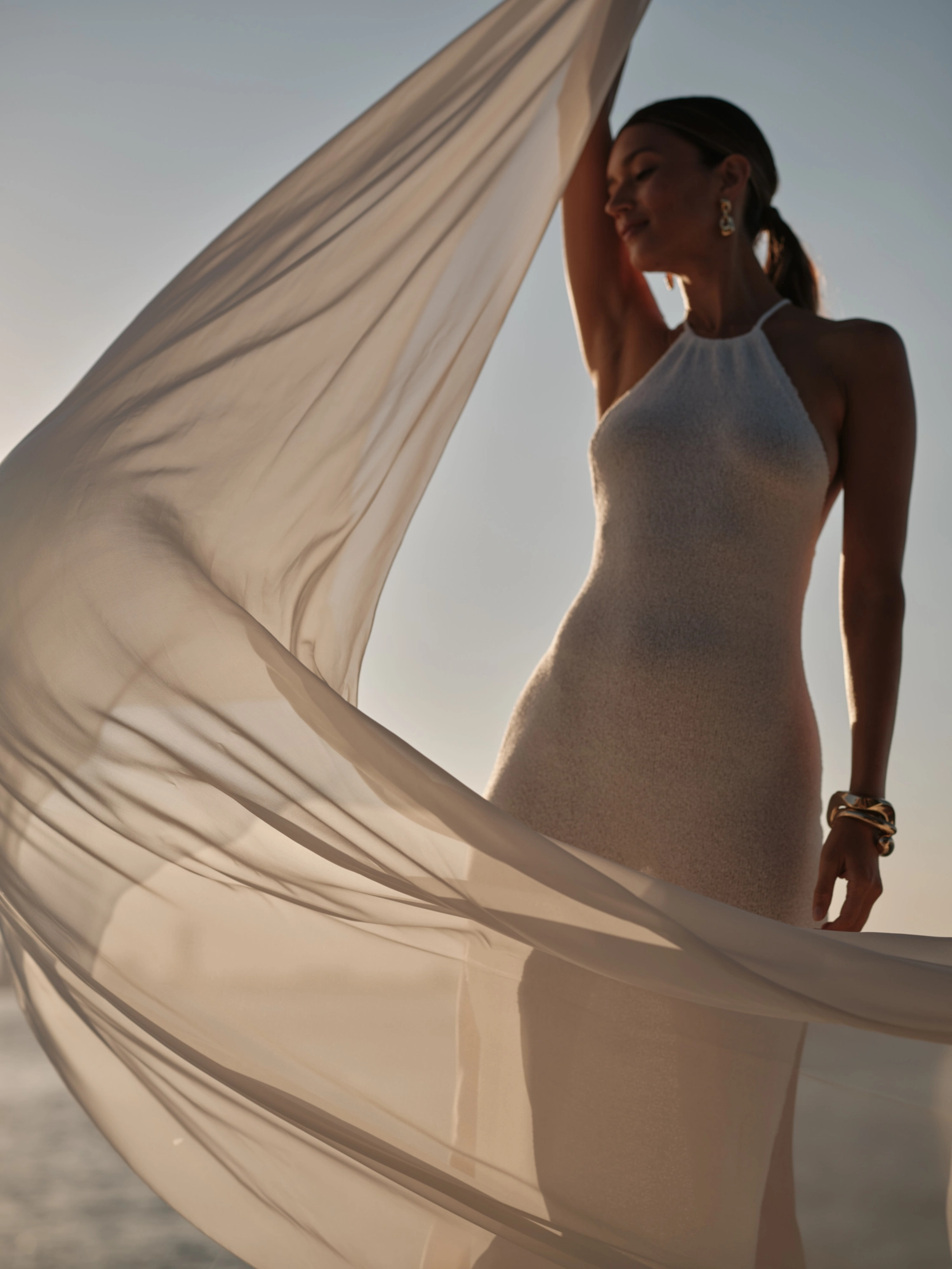 LILLI model in white dress holding flowing fabric against the light blue sky.